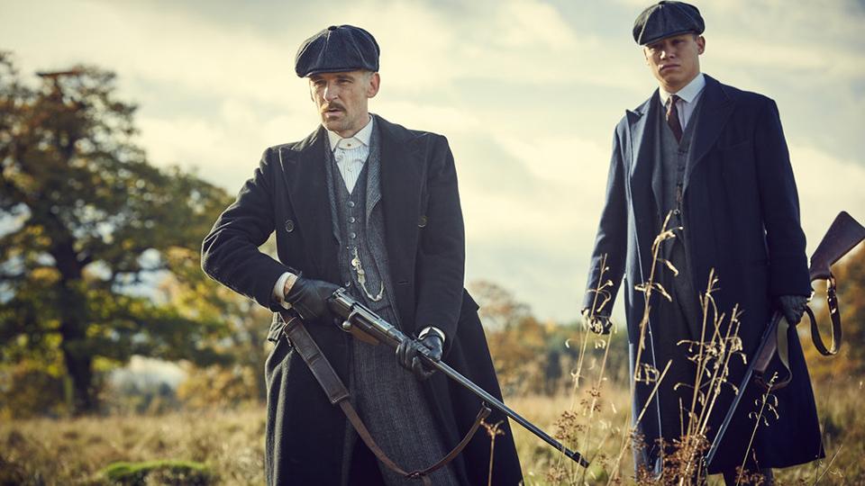 Image of Peaky Blinders characters Arthur Shelby and Michael Gray in a field, dressed in grey suits and coats, carrying shotguns