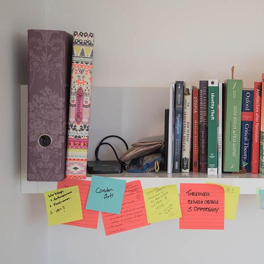 A student's bookcase on a white wall with brightly coloured folders and books and post-its hanging off it.