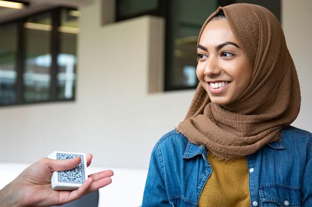 A student wearing a brown hijab and denim shirt smiles as they are shown a magic trick via a pack of cards