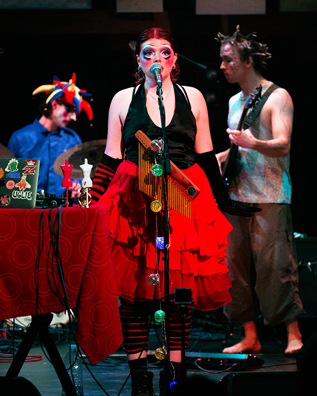 A woman singing on stage with theatrical make-up. Behind her stands another musician in a jester hat and a man with spiked hair playing guitar.