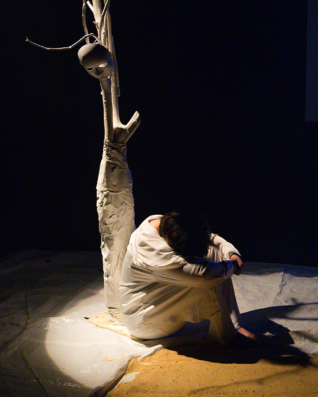A person sits on a dimly lit stage, curled into a ball. Behind them stands a white tree-like structure with a theatrical mask hanging from a branch.