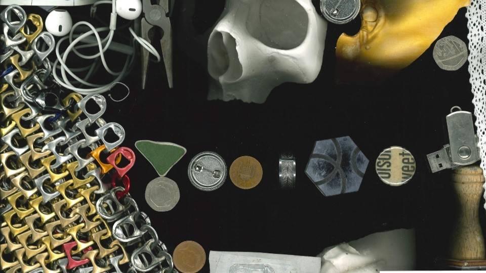 a selection of odd objects arranged on a black background, including a skull, coins, a badge, a piece of ceramic, some pliers, a pair of headphones 