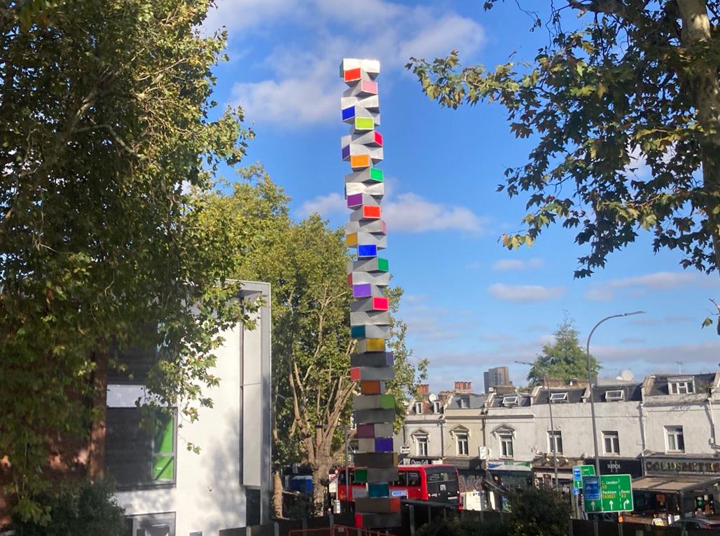 A tall multicoloured sculpture framed by buildings and trees