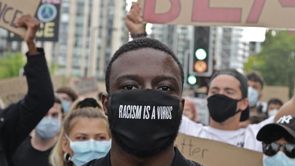 Photograph shows a young black man at a protest with a face mask which has the words 'racism is a virus' on it