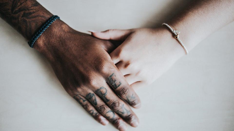 A photograph of a darker masculine tattooed hand and a pale female hand touching
