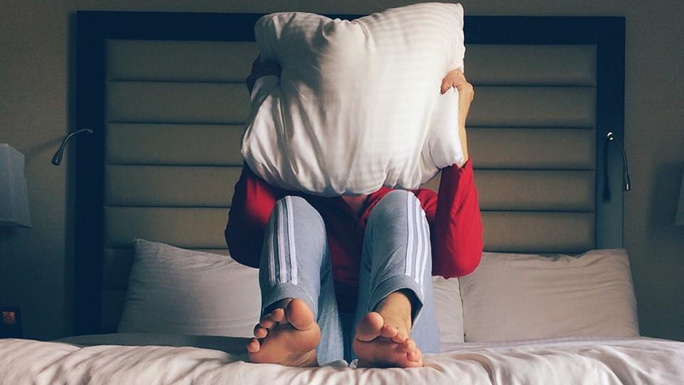 A young man sits on a bed with a white pillow over his head