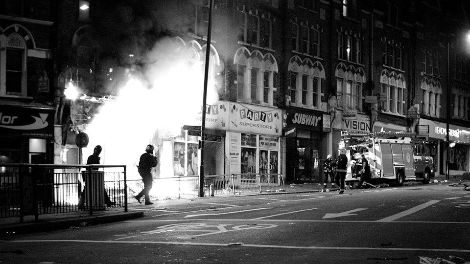 A black and white photo of a road with a row of shops. One is on fire, two people are in front of it