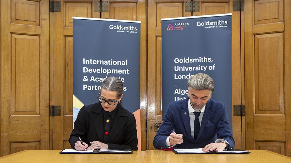 Image shows a woman (left) and a man in formal clothing sitting at a table signing documents in front of two branded banners and some wooden doors