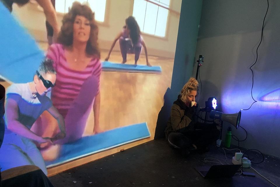 Artist sitting on floor in front of screen showing exercise studio