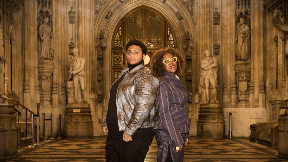 Young man in grey jacket (left) back to back with woman (right) in purple suit