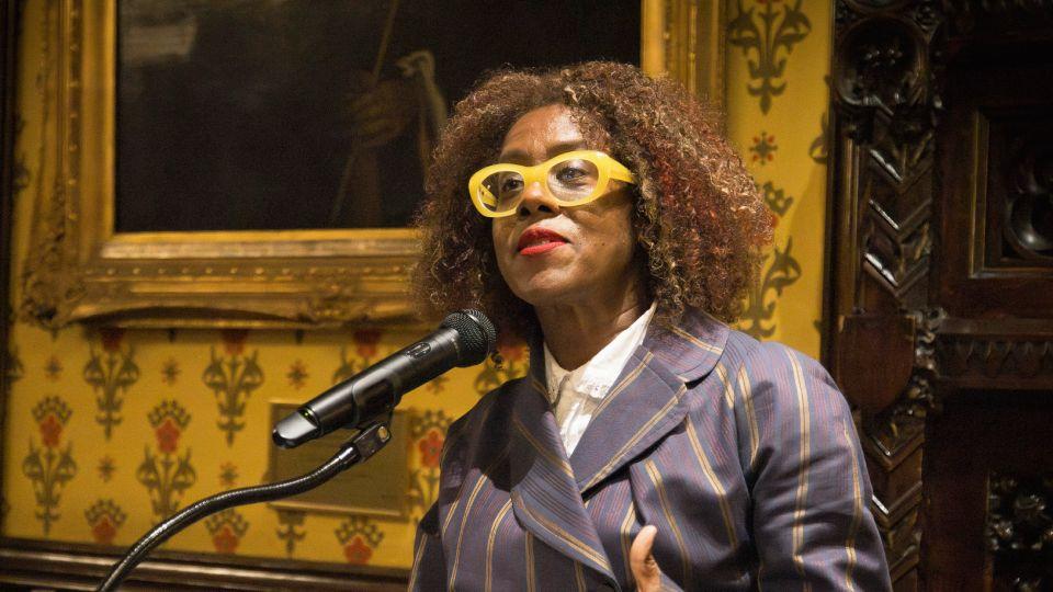 Female in striped suit, with yellow framed glasses addresses guests