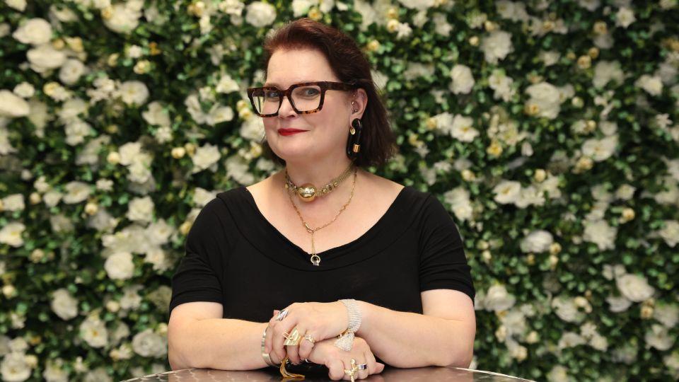 woman in glasses, torso and head, smiles, arms crossed. Flora background. 