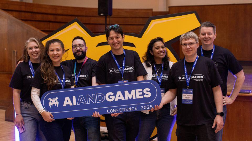 The conference organisers wearing matching T shirts and holding a sign with the logo and saying AI and Games conference