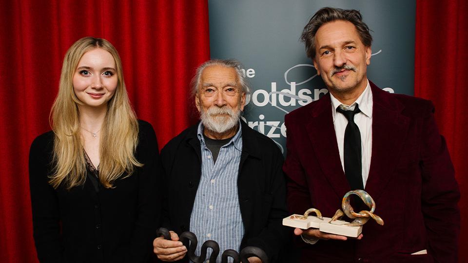 Young Writer competition winner Katie Parkinson with Goldsmiths Prize winners M. John Harrison (2020) and C.D. Rose (2025) holding their trophies in front of red curtains and a banner which says The Goldsmiths Prize