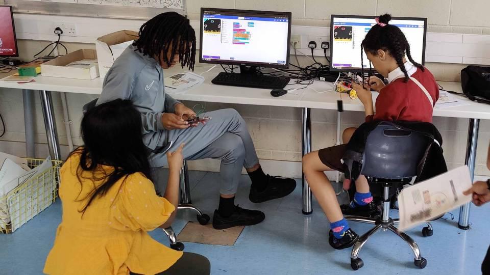 Female in yellow seated on floor works with two pupils who are seated at desks