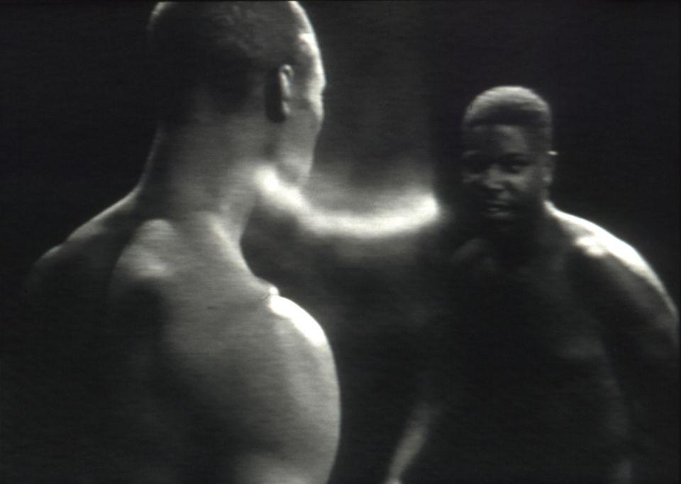 A still from a black and white film of two topless men standing opposite each other. 