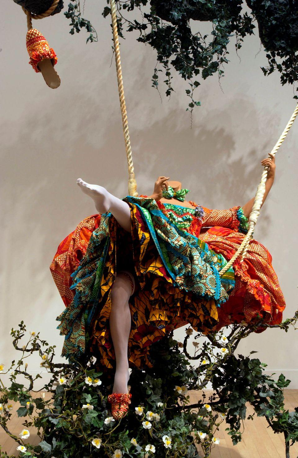 A sculpture of a headless, life-size mannequin on a rope swing wearing a dress made from colourful Dutch wax printed cotton textile surrounded by faux foliage. 