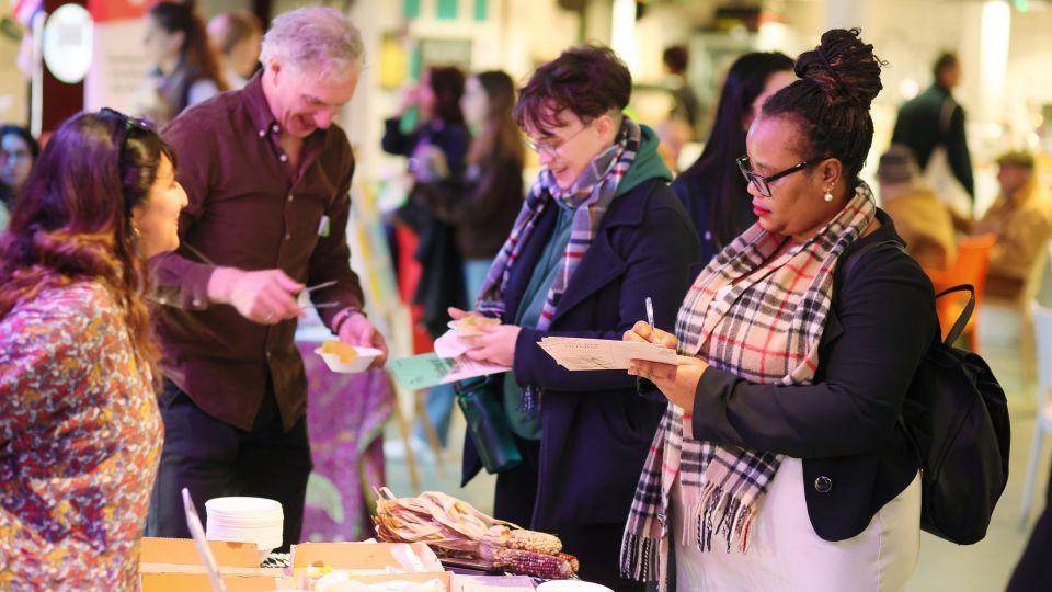 A group of people by a table. A woman behind the table talks to a woman who is holding a notepad and pen. Two people in the background holding food.