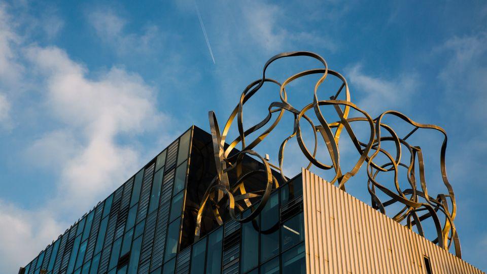 The grey Ben Pimlott building with its metal squiggle sculpture on top against a blue sky