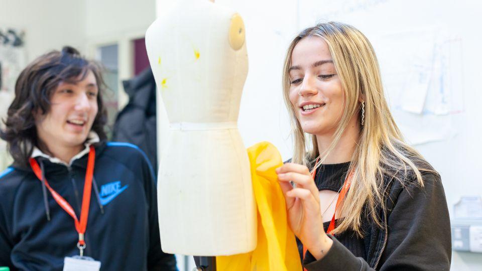 Two people, one is holding yellow fabric up against a dressmaking mannequin 