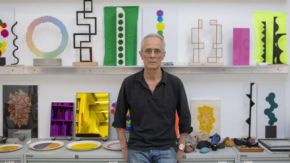 A portrait of artist David Batchelor wearing a black polo shirt and glasses leaning on a bench covered in artworks and with a shelf behind him with models and small sculptures.