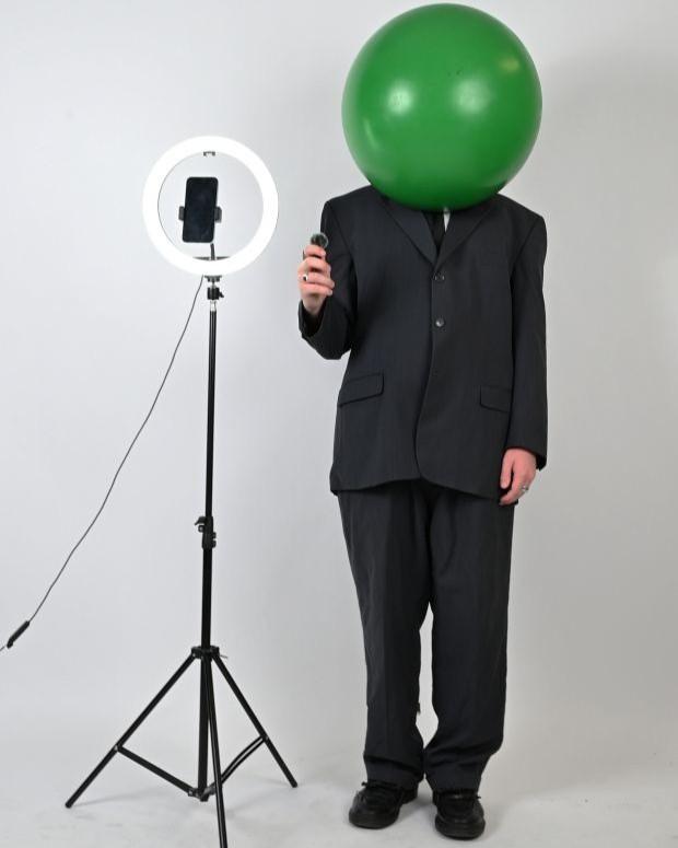 A man in a black suit but over his head he is wearing a green orb. There is a ring light on a stand and he is holding a small microphone. 