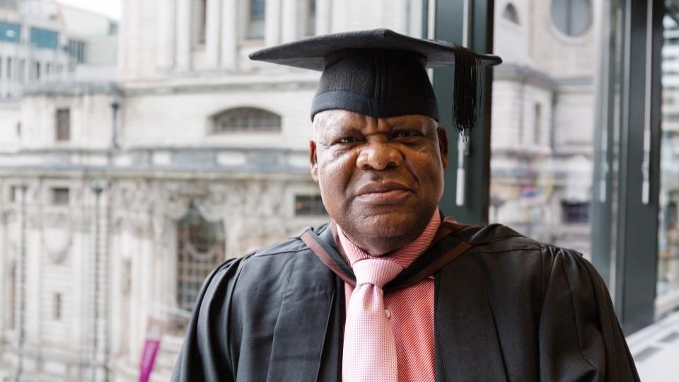 Chris wearing a cap and gown over a stripy red and white shirt and tie, pictured in front of a window 