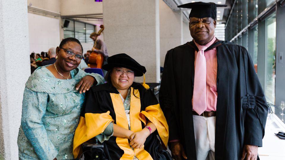 A woman on the left wearing a pale blue outfit, a woman in the centre wearing academic robes with yellow details and a cap, and a man on the right wearing a graduation cap and academic robes over a red striped shirt