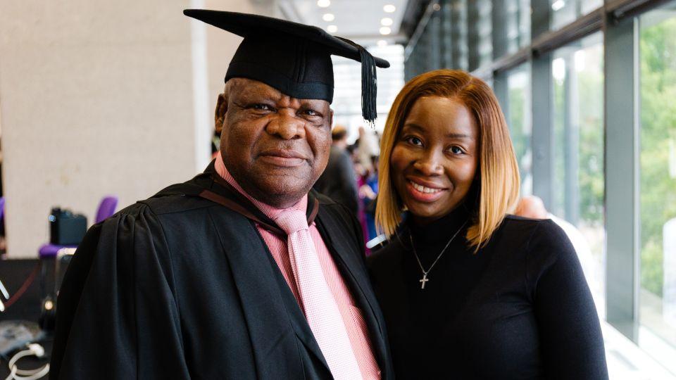 A man in a graduation cap and gown over a red striped shirt smiles for a photo with a woman wearing a black top