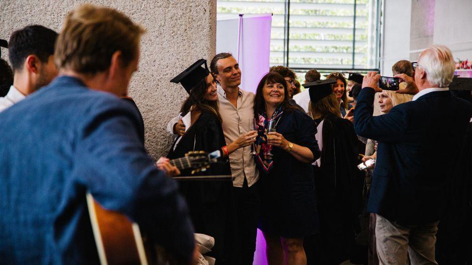 In the foreground, a man playing guitar seen side on. In the background, three people pose for a photo toasting prosecco. 
