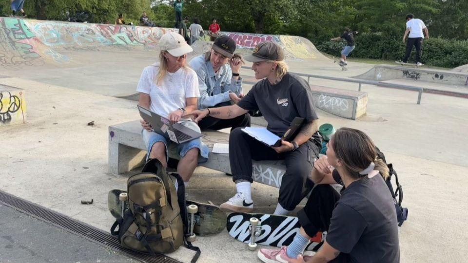 Four people are sat around a concrete bench at a skatepark gathered round a laptop having a conversation, and there are people skateboarding in the background