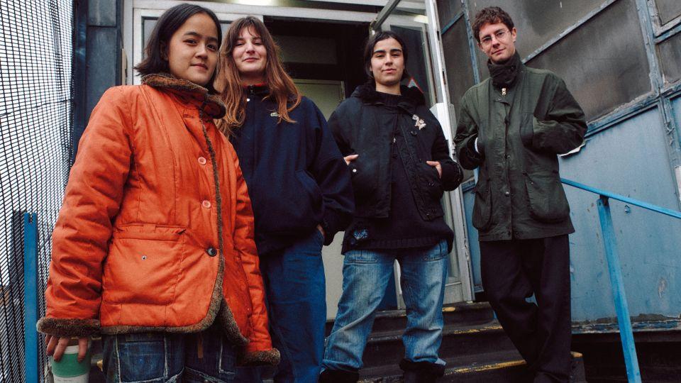 The four graduates wearing their coats stand for a photo on steps outside by a doorway