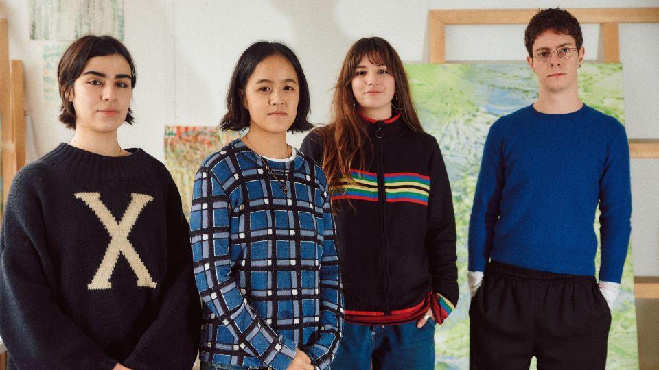 The four graduates stand in a row in a studio space, with large paintings and wooden frames visible behind them