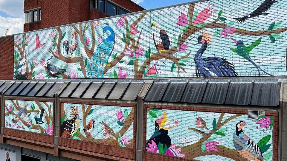 A large mural painted on a brick wall, with a blue background, branches, pink flowers and lots of different types of birds