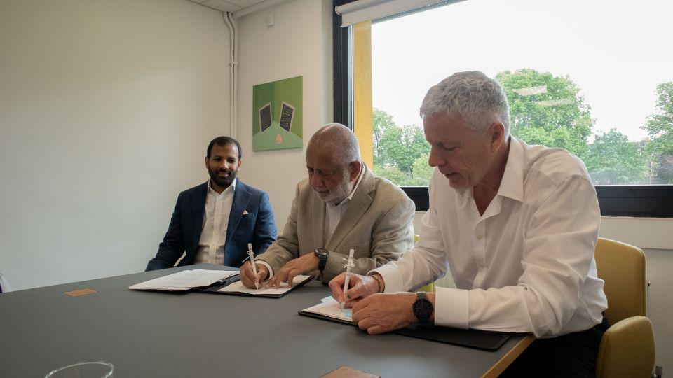 Three men are sitting at a table, two are holding pens and signing papers