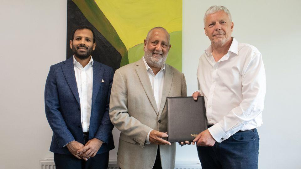Three men post for a photograph in front of a painnting, with two of the men holding a folder with the Goldsmiths logo.