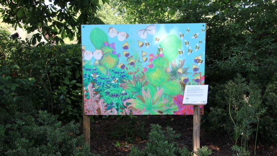 A board surrounding by trees. It shows a colourful artwork with plants, butterflies and bees. 