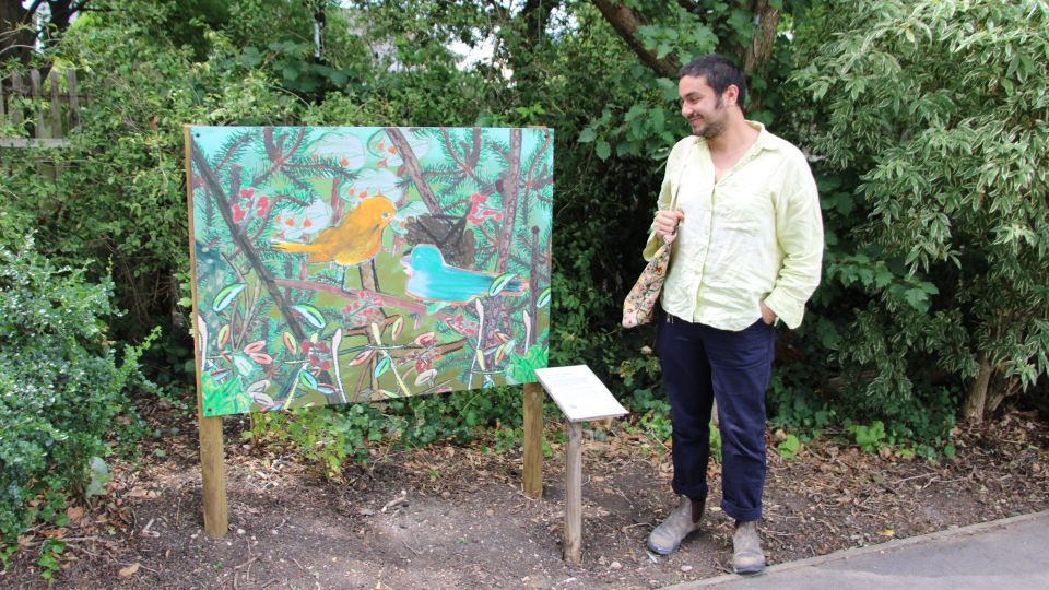 A board with a colourful painting showing an orange bird and branches, in front of some trees, and a man in a yellow shirt and dark blue trousers. 