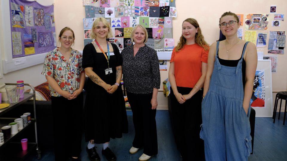 Five women smile for a photograph in front of a wall decorated with displays of creative artworks