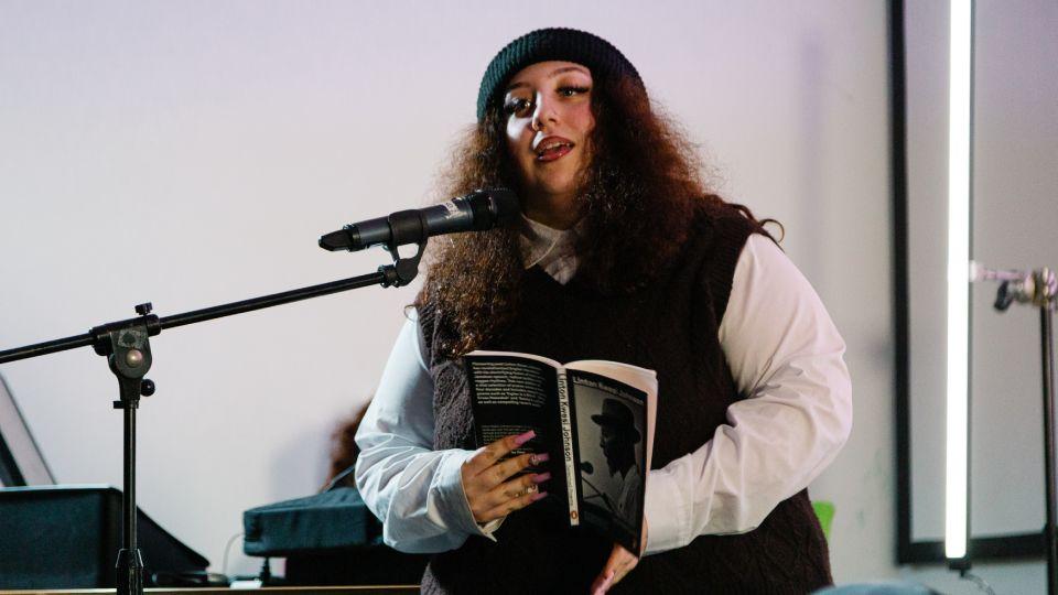 Young woman, long hair wearing beanie cap, talks into microphone on stand