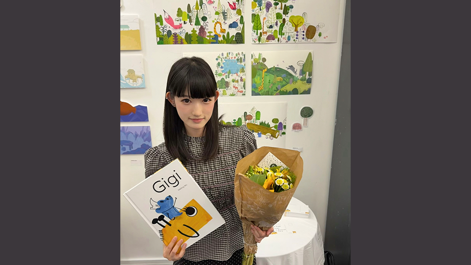 Meiling is holding her book, Gigi, and a bouquet of flowers and illustrations from her book are displayed on the wall behind her
