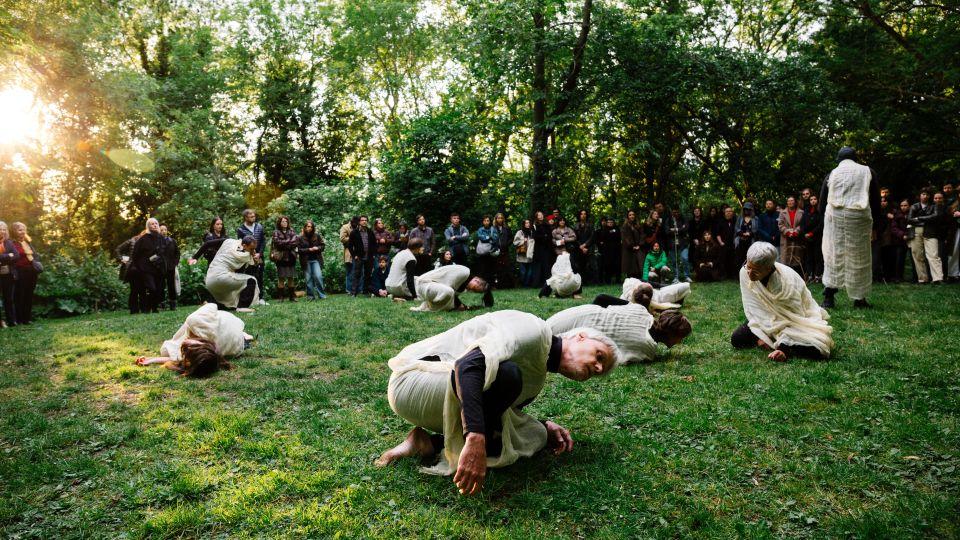 Dancers wearing white shrouds over black clothing performing 'Wither and Bloom' in the glade
