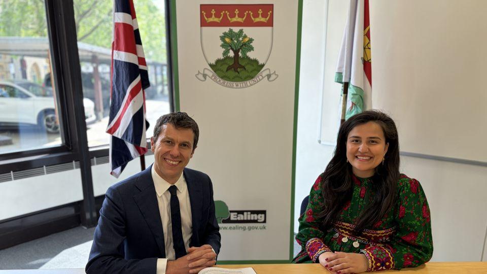 Tony Clements wearing a suit and tie and Rabia Nasimi wearing a green and pink dress sat at a desk in front of a sign with the Ealing crest