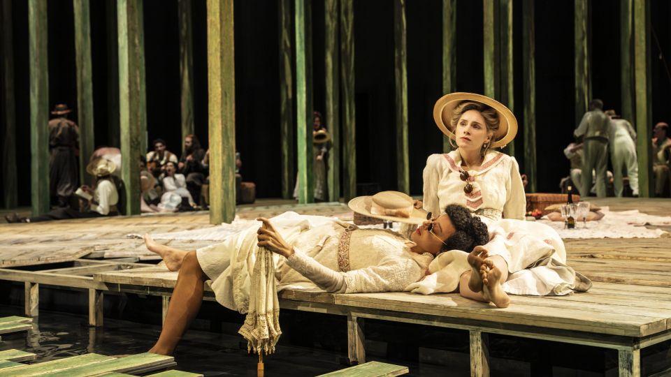 A woman wearing a hat sat on wooden decking with another woman lying down. 