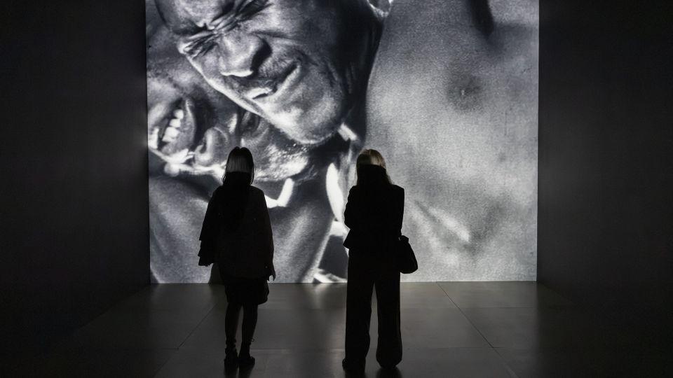 Two people standing in front of a wall where a black and white film is being projected showing two men wrestling. 
