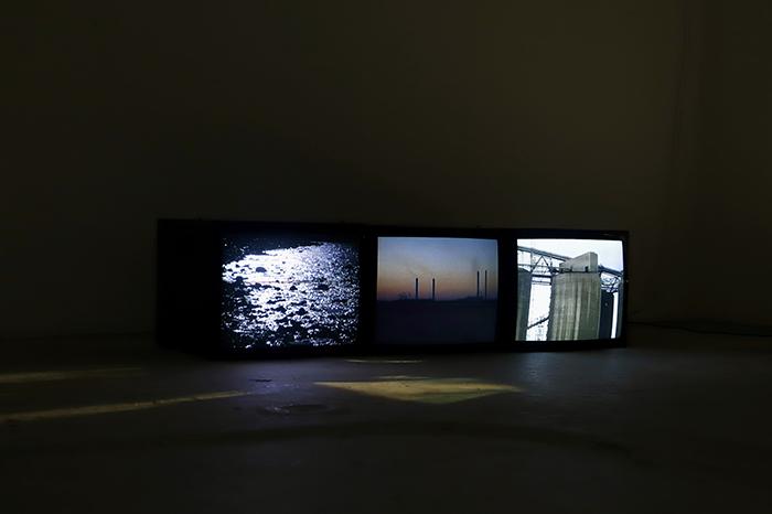 Three television screens in the dark showing Kuwait's industrialised northern coastline