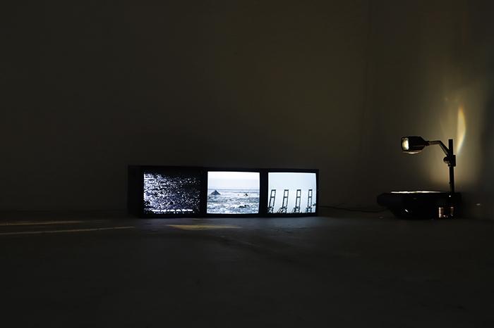 Three television screens in a dark room, showing Kuwait's industrialised northern coastline