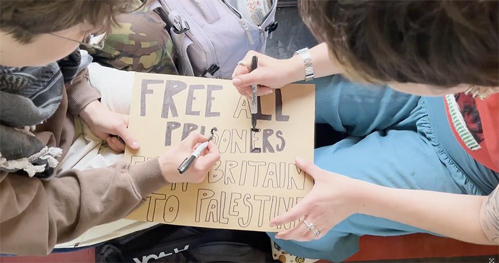 Two people sit close together, using markers to write a protest message on a cardboard sign.