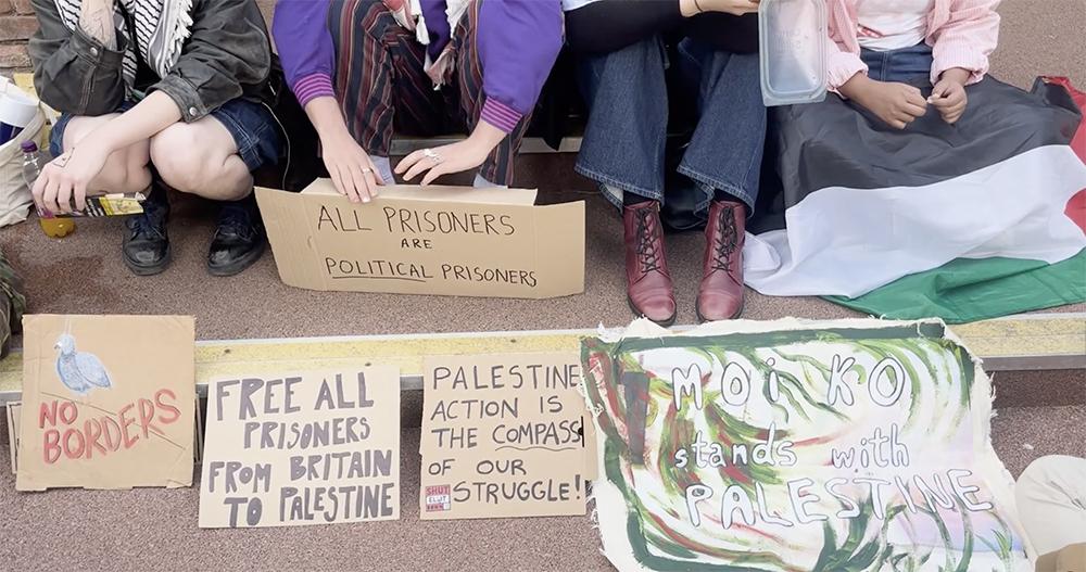 A group of people sit on the ground with cardboard protest signs arranged in front of them. The signs read “All prisoners are political prisoners,” “Free all prisoners from Britain to Palestine,” “Palestine action is the compass of our struggle,” and “No borders.”