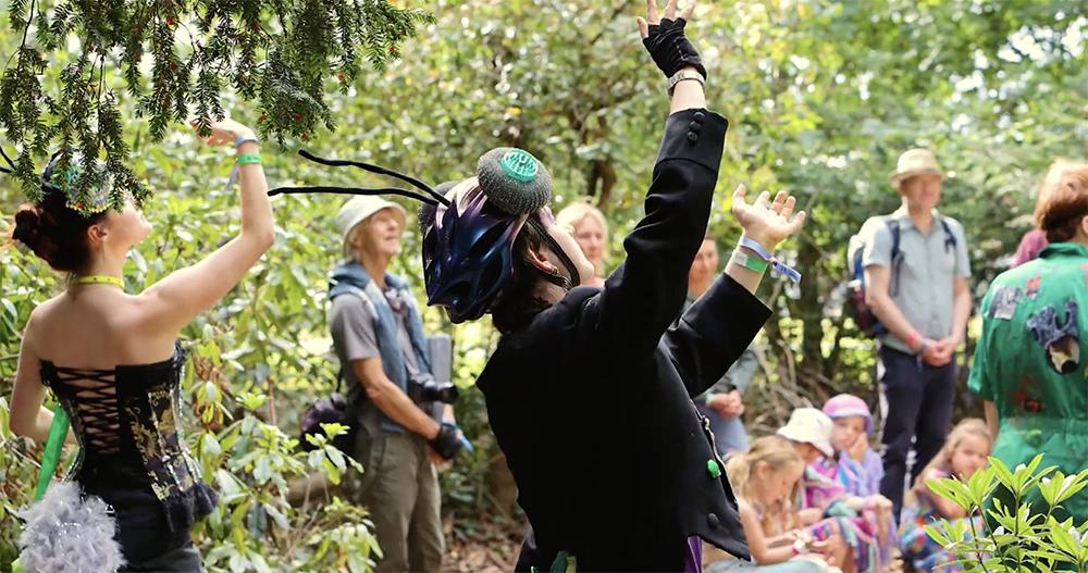 People gather in a wooded outdoor setting as two performers in costume raise their arms toward the trees, appearing to dance or gesture expressively. Onlookers of various ages stand and sit nearby among greenery, watching the performance.